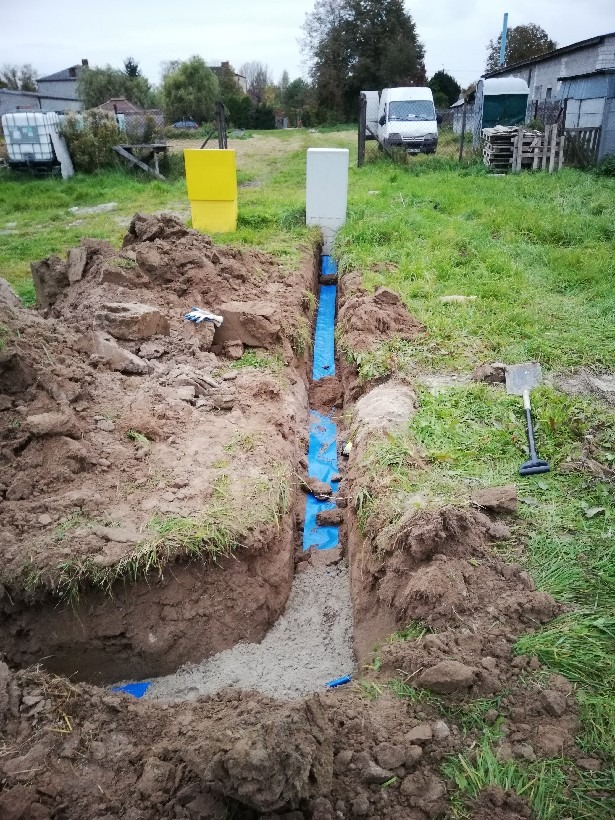 Wykop w ziemi z ułożonymi niebieskimi rurami, częściowo zalany betonem, widoczne skrzynki elektryczne i łopata w tle.