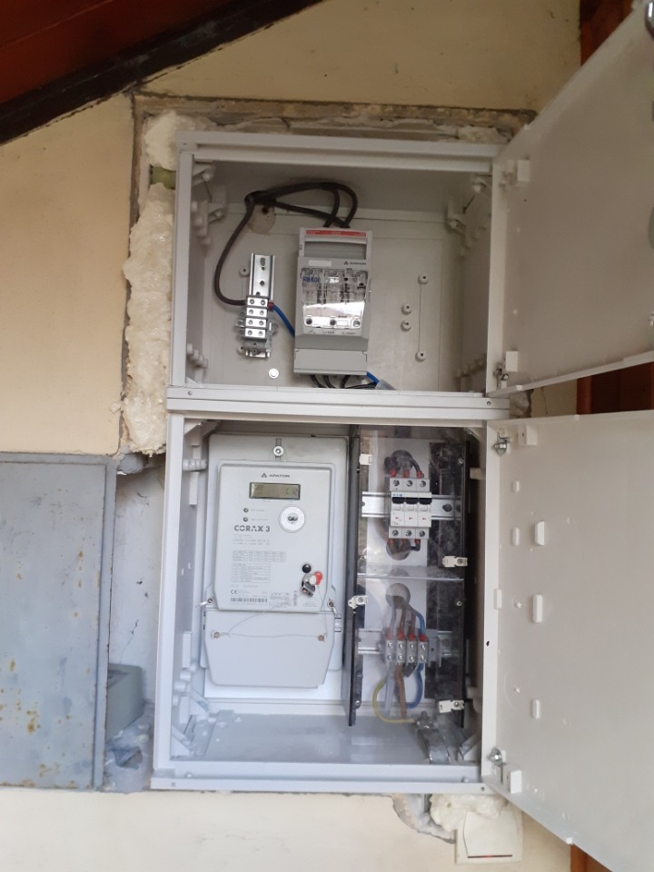Otwarta, dwupoziomowa skrzynka elektryczna z dwoma licznikami energii, zabezpieczeniami i przewodami, zamontowana na nierównej ścianie z widocznymi uszkodzeniami i pianką montażową.