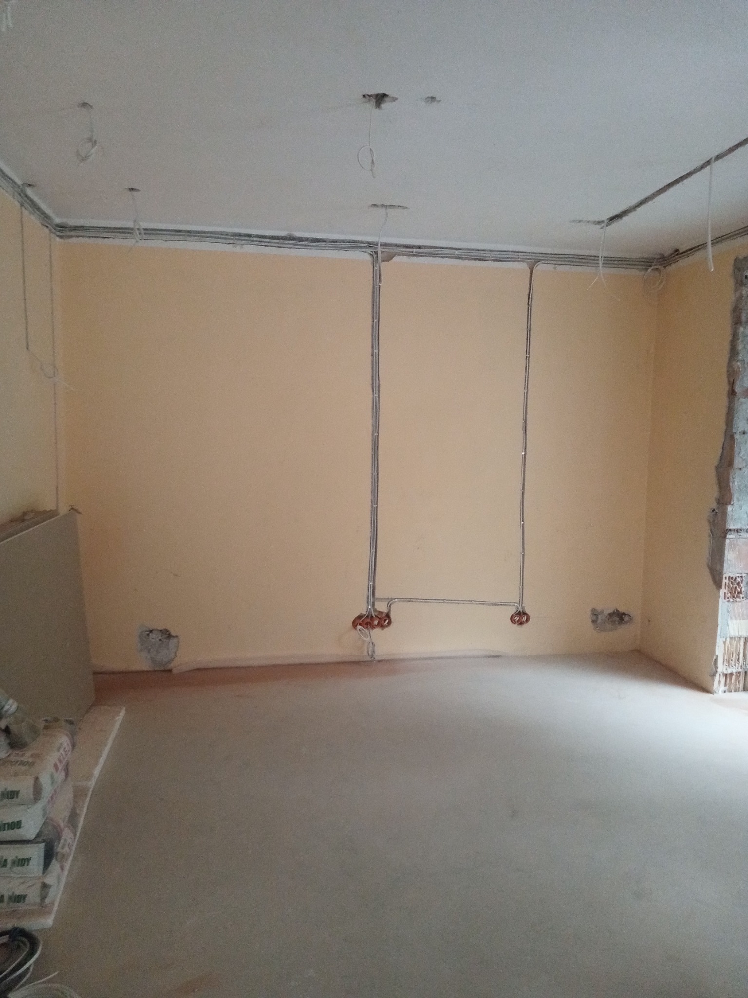 Wnętrze w trakcie remontu z poprowadzonymi przewodami elektrycznymi po ścianach i suficie, widoczne puszki instalacyjne, worki z zaprawą na podłodze.