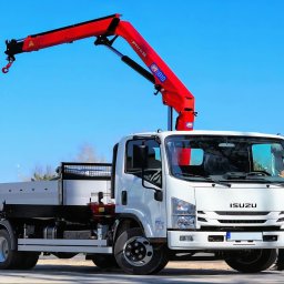 HOŁDA - Biała ciężarówka Isuzu z czerwonym dźwigiem hydraulicznym na tle błękitnego nieba, zaparkowana na placu budowy z paletami w tle. Widok z przodu.