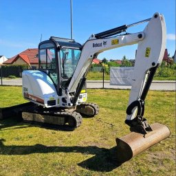 HOŁDA - Biała mini koparka Bobcat 334 na trawie, z łyżką skierowaną w dół, na tle domów i błękitnego nieba. Widok z boku, maszyna gotowa do pracy.