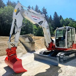 HOŁDA - Biało-czerwona koparka Takeuchi na placu budowy z hałdami piasku w tle, gotowa do pracy. Widoczny lemiesz i łyżka koparki.