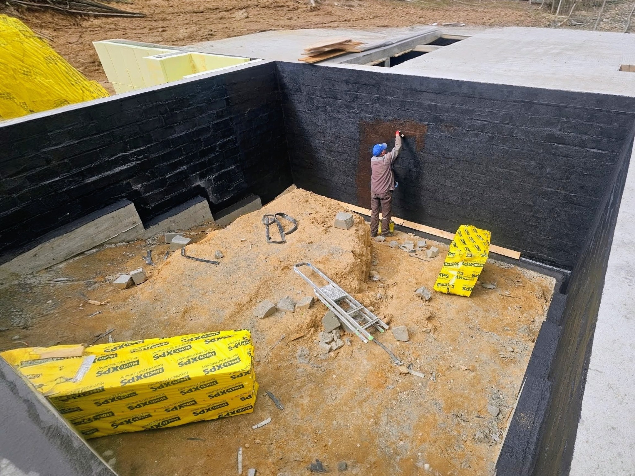Hydroizolacja fundamentów: pracownik maluje czarną powłoką ścianę wykopu. Widoczne materiały izolacyjne XPS w żółtym opakowaniu i drabina.