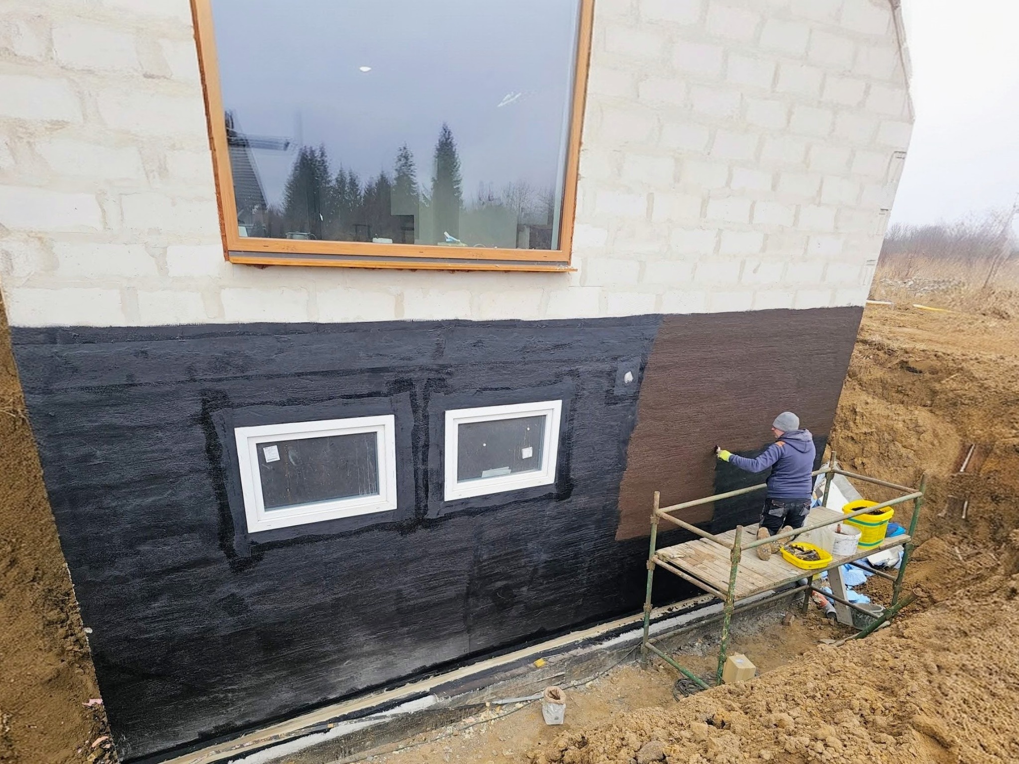 Fundament budynku z nałożoną czarną hydroizolacją, pracownik na rusztowaniu aplikuje preparat. Widoczne okna i wykop wokół fundamentu.