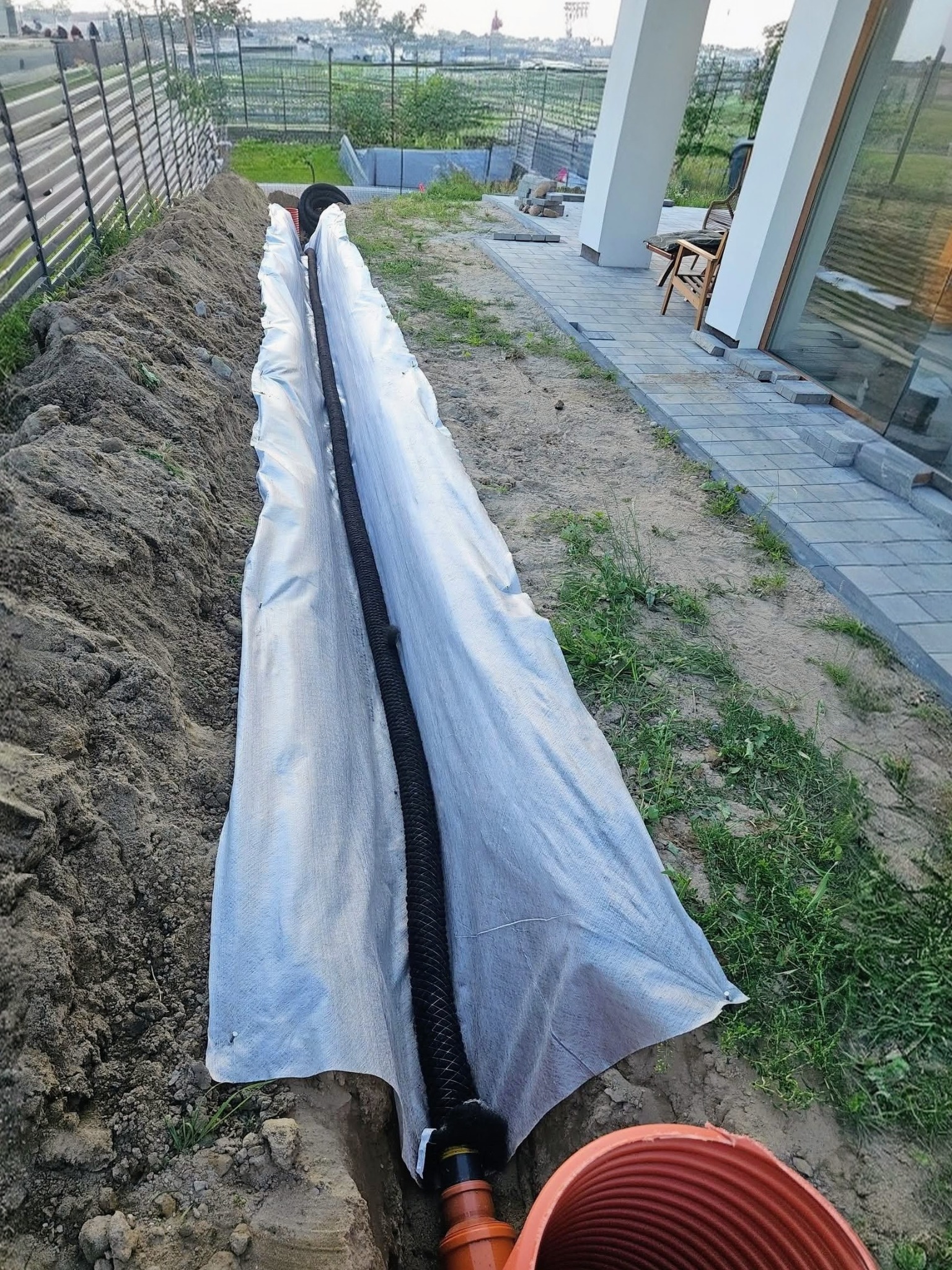 Wykop z rurą drenażową w geowłókninie, widok z góry. Rura częściowo zakopana, obok kostka brukowa i trawa. Elementy domu w tle.