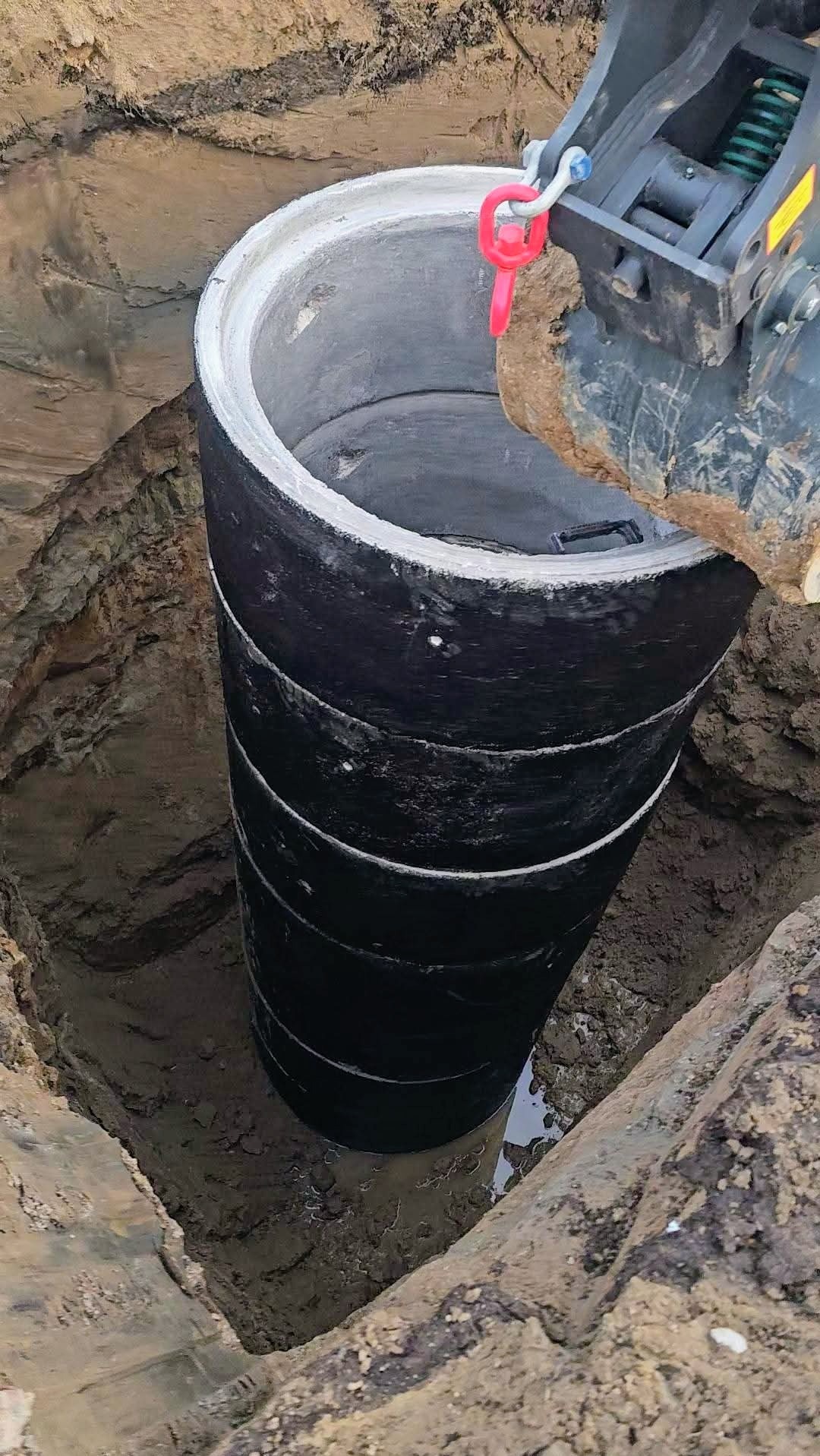 Montaż czarnego, betonowego szamba w wykopie ziemnym za pomocą koparki. Widoczne łączenia segmentów szamba i czerwony uchwyt mocujący.