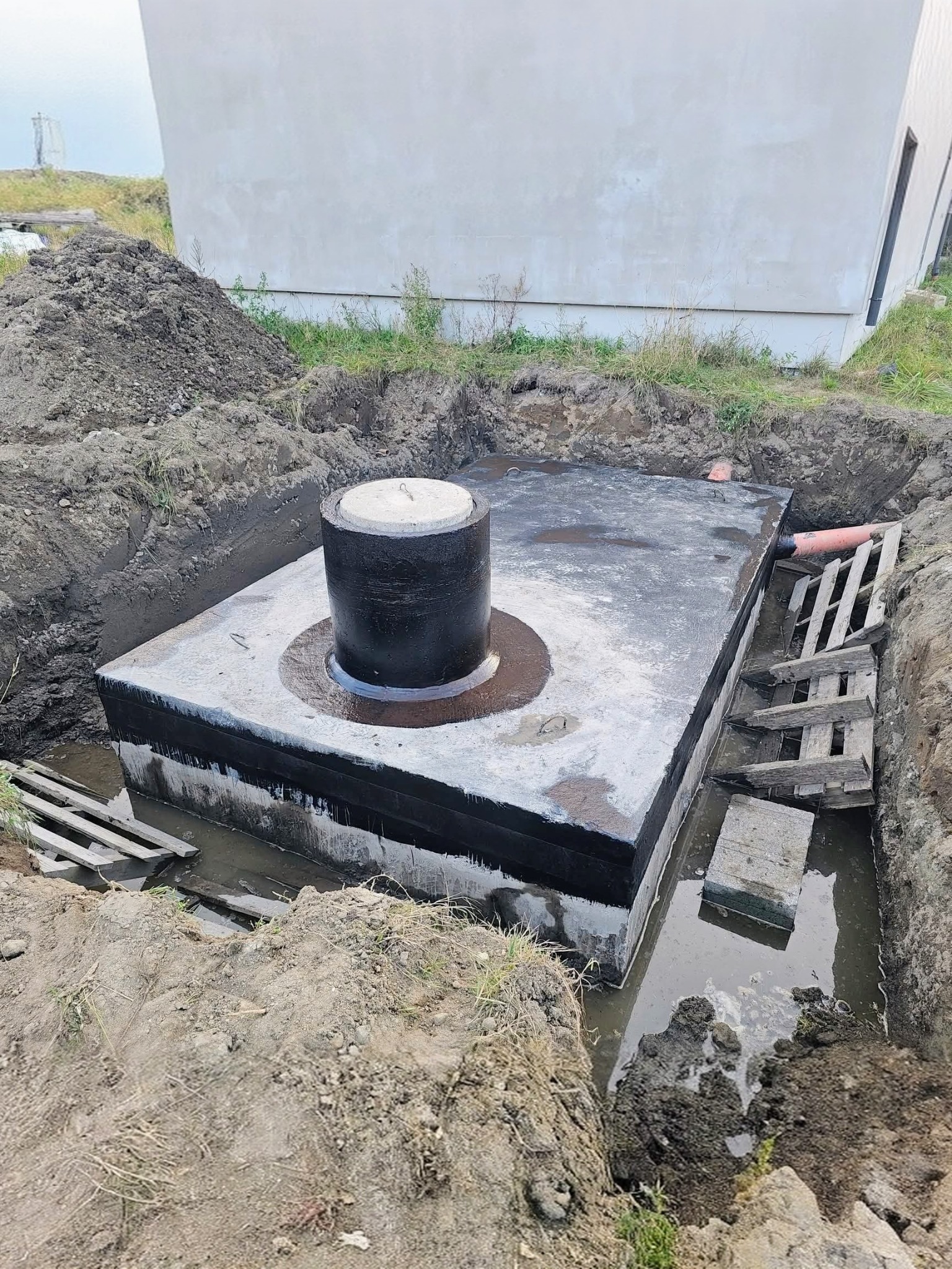 Betonowy szambo w wykopie, zabezpieczony izolacją, z widoczną rurą odpływową i drewnianymi elementami szalunku. Częściowo zalany wodą, ziemia wokół.