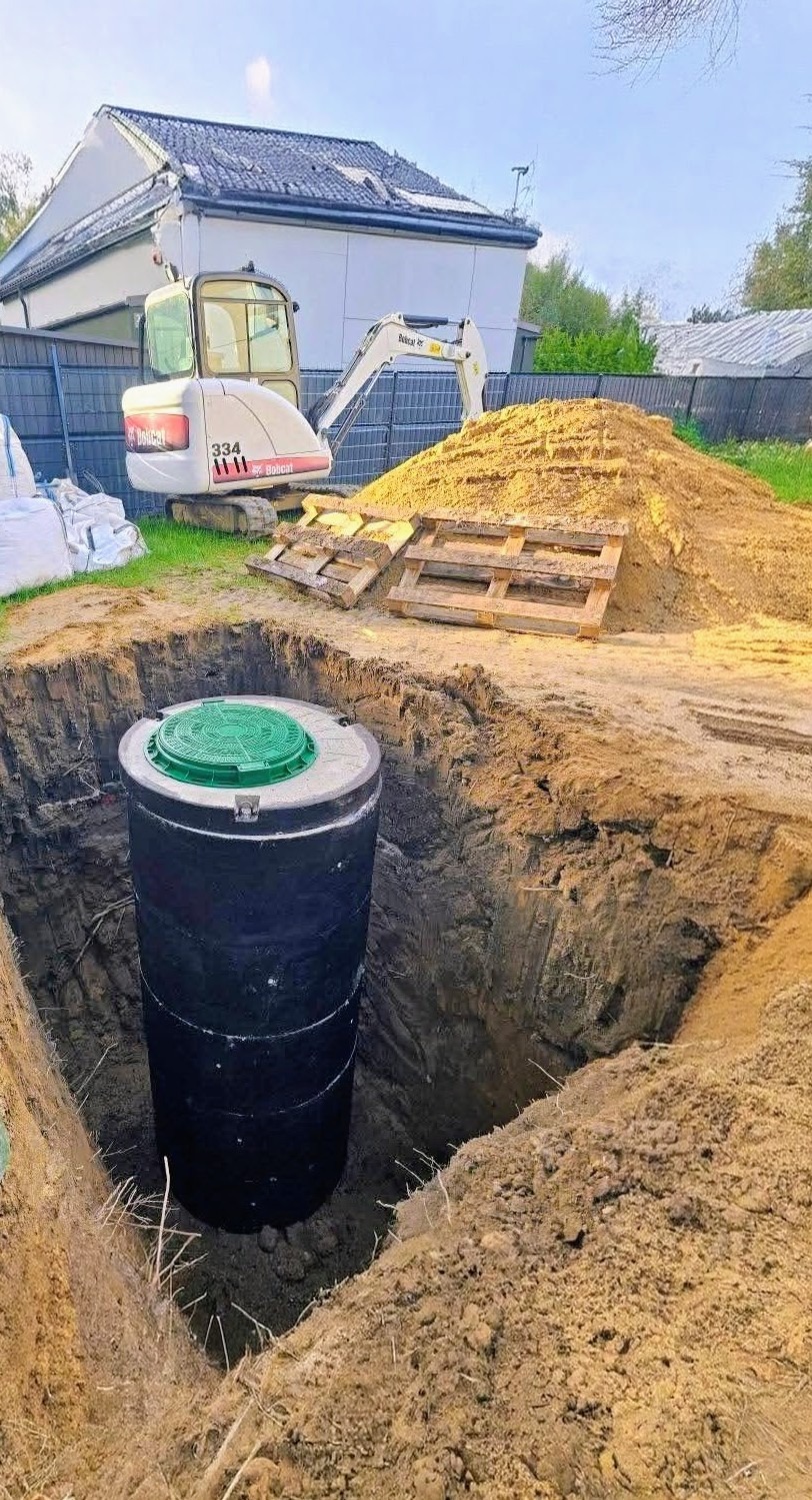Czarny zbiornik z zieloną pokrywą w wykopie, w tle minikoparka Bobcat i pryzma ziemi. Widok z góry na dół, słoneczny dzień.