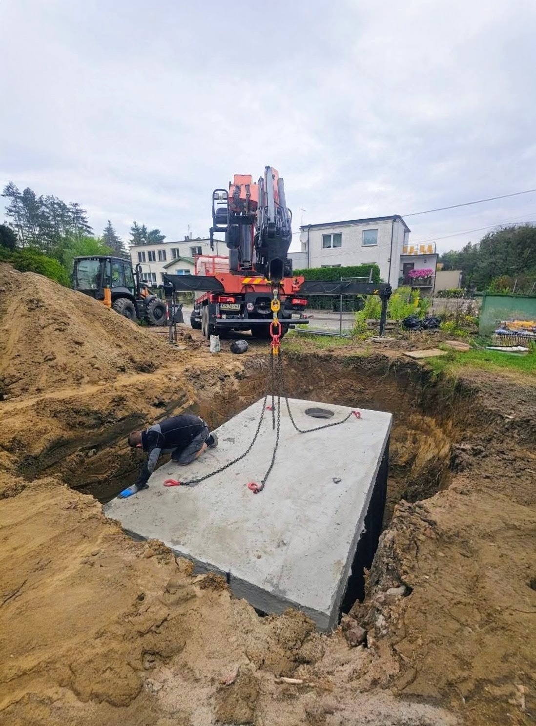 Montaż betonowego szamba w wykopie za pomocą dźwigu. Pracownik w wykopie, w tle budynki mieszkalne i sprzęt budowlany. Szambo z otworem na górze.