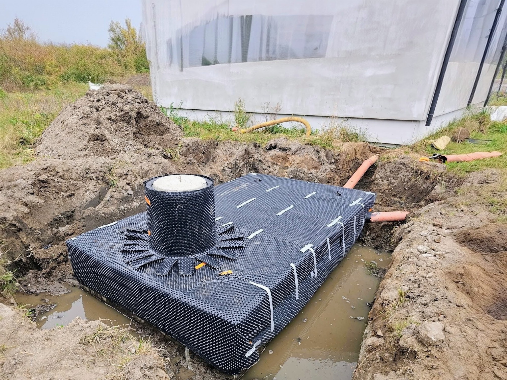 Montaż szamba betonowego w wykopie, zabezpieczonego folią kubełkową, z widocznymi rurami odpływowymi i stertą ziemi w tle. Częściowo zalane wodą.