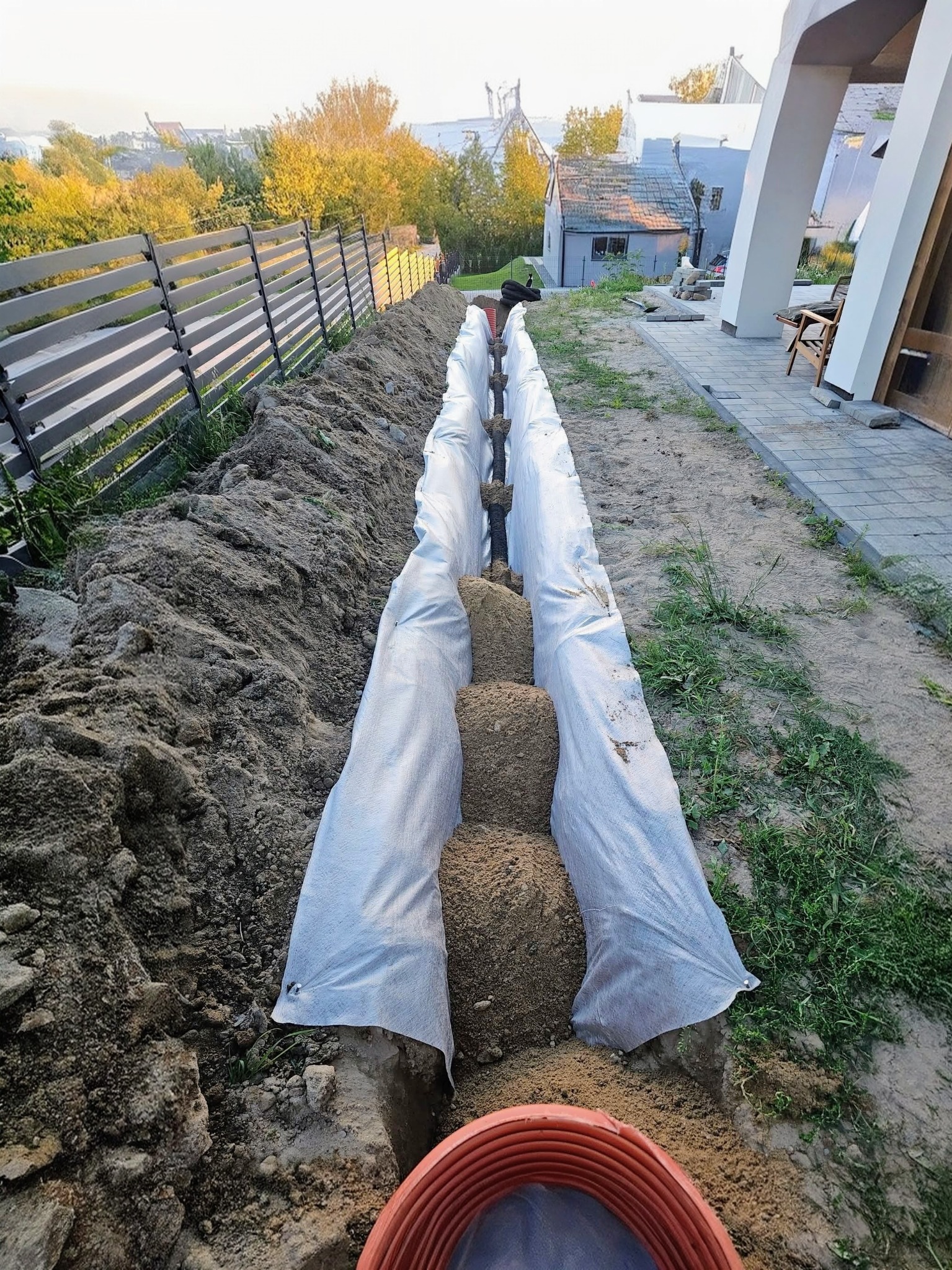 Wykop z rurami drenażowymi owiniętymi geowłókniną i zasypanymi piaskiem, widok z góry. Prace ziemne w ogrodzie, obok kostka brukowa i płot.