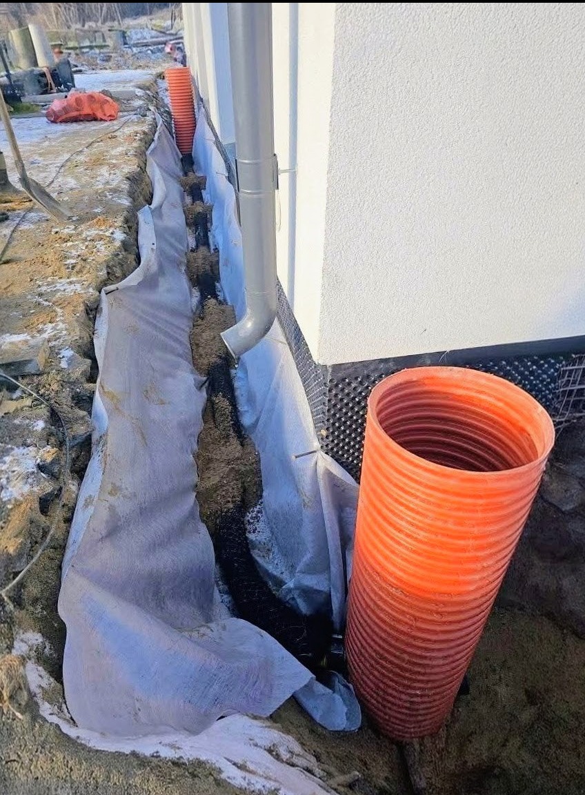 Drenaż opaskowy budynku w trakcie realizacji. Widoczna rura drenarska w wykopie, zabezpieczona geowłókniną. Rura spustowa i fragment elewacji budynku.