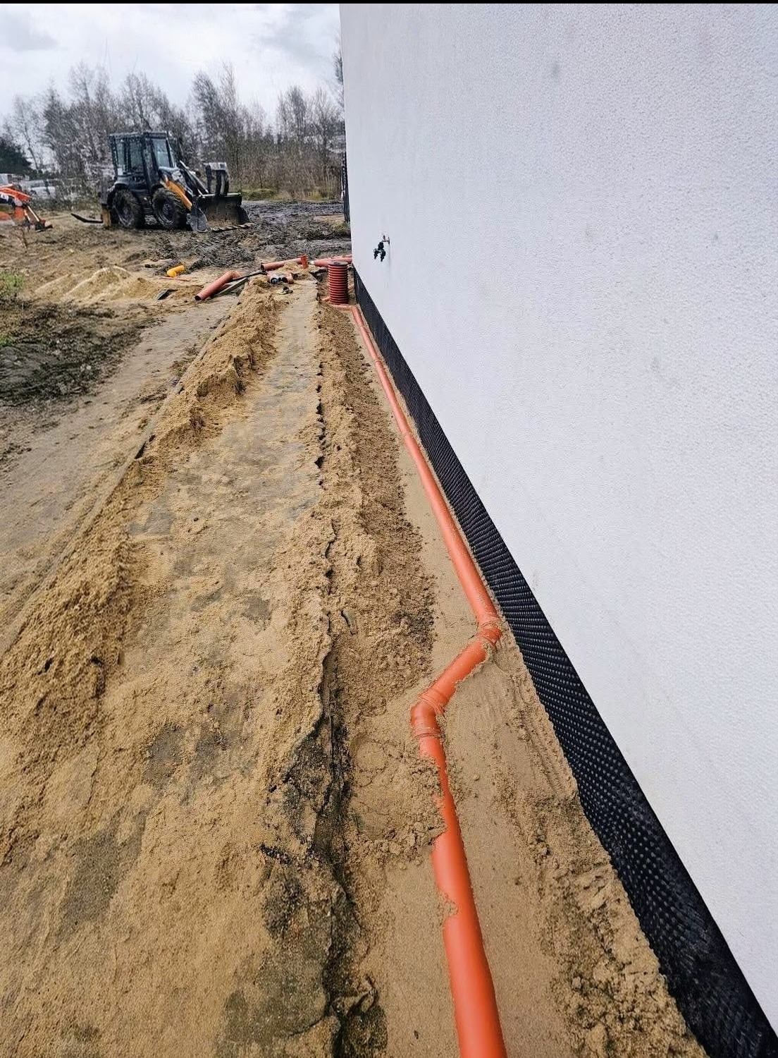 Instalacja drenażowa z rur PVC przy fundamencie budynku, widoczna czarna folia izolacyjna i wykop. W tle koparka.