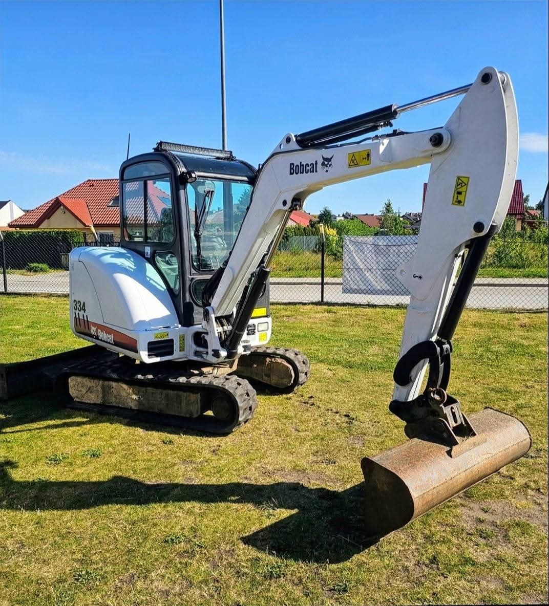 Biała mini koparka Bobcat 334 na trawie, z łyżką skierowaną w dół, na tle domów i błękitnego nieba. Widok z boku, maszyna gotowa do pracy.