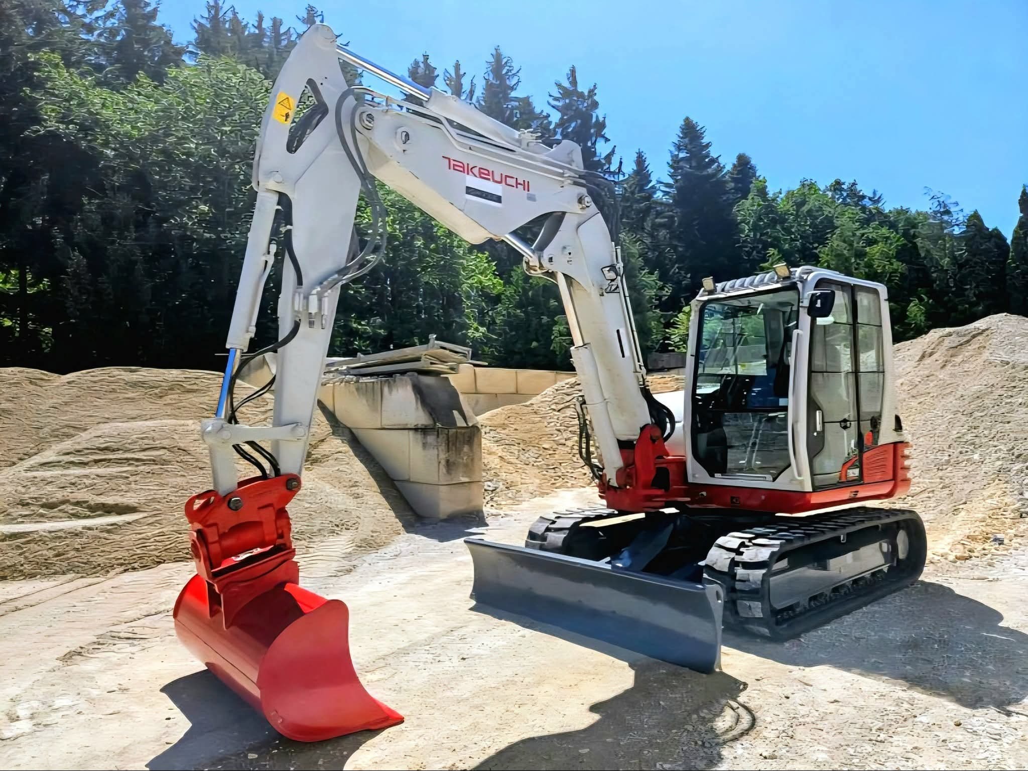 Biało-czerwona koparka Takeuchi na placu budowy z hałdami piasku w tle, gotowa do pracy. Widoczny lemiesz i łyżka koparki.