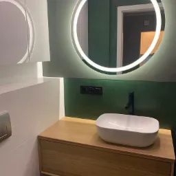 Nowoczesna łazienka z okrągłym lustrem LED, umywalką nablatową na drewnianej szafce i czarną baterią, zielona ściana w tle.