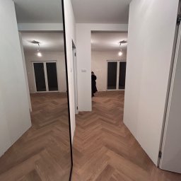 K&K Remonty Spółka z ograniczona odpowiedzialnoscia - Parkiet w jodełkę w jasnym korytarzu, odbicie w lustrze. Widoczne białe ściany i drzwi. Minimalistyczny design wnętrza, ciepłe oświetlenie.