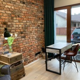 K&K Remonty Spółka z ograniczona odpowiedzialnoscia - Loftowe wnętrze z ceglaną ścianą, drewnianą podłogą i designerskimi meblami. Widoczny stolik, krzesło i komoda w stylu vintage. Z okna widok na dom i samochód.