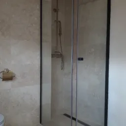 Kabina prysznicowa z czarnymi profilami i deszczownicą w stylu retro, wykończona płytkami imitującymi trawertyn.