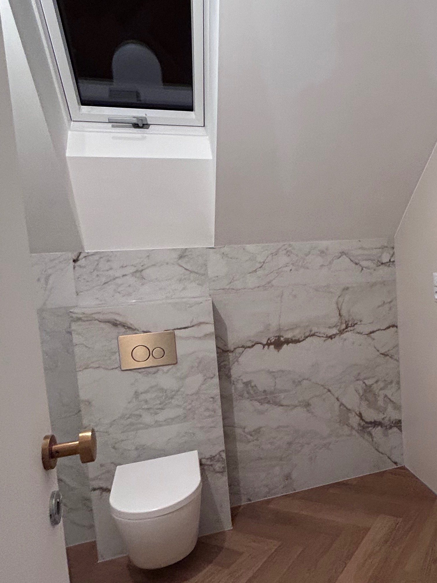 Wykończona łazienka w Milanówku: biała wisząca toaleta z złotym przyciskiem spłukiwania na tle marmurowych płytek. Podłoga wyłożona parkietem w jodełkę, okno dachowe.