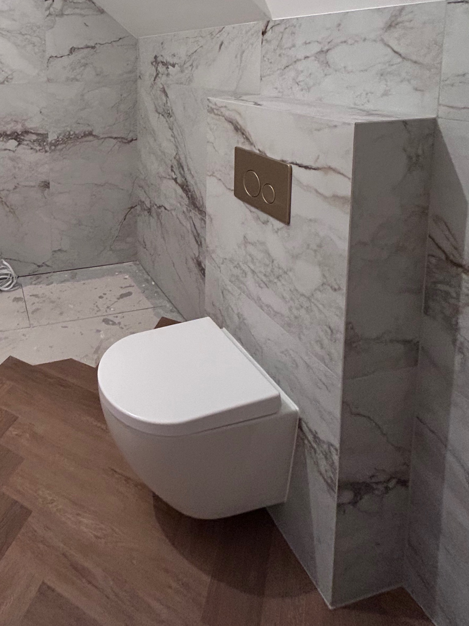 Nowoczesna łazienka z podwieszaną toaletą, wykończona płytkami imitującymi marmur i drewnianą podłogą w jodełkę. Widoczny złoty przycisk spłuczki.