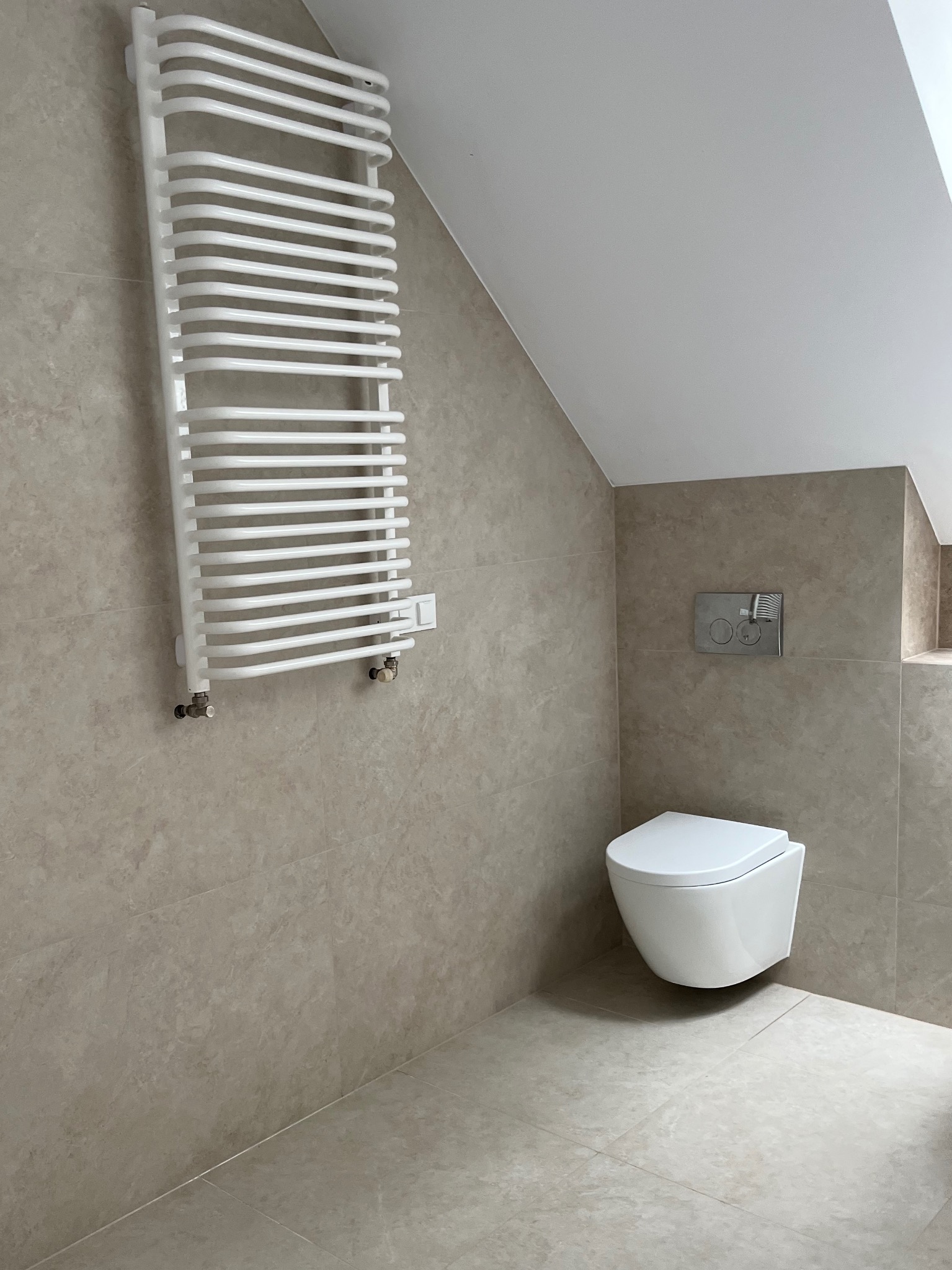 Beżowa łazienka na poddaszu z wiszącą toaletą i grzejnikiem drabinkowym. Płytki imitujące kamień, minimalistyczny styl, skosy sufitowe.