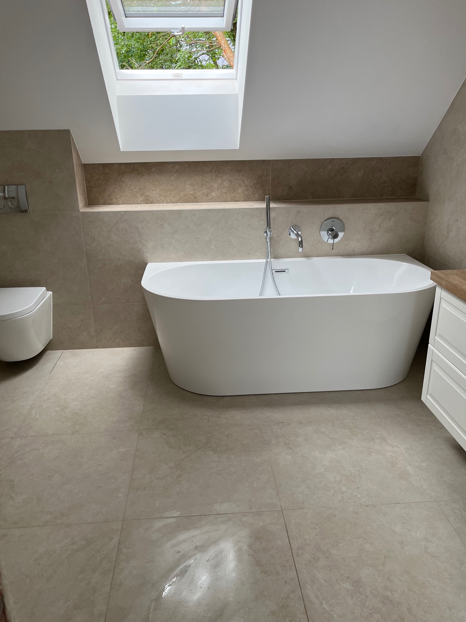 Nowoczesna łazienka na poddaszu z wolnostojącą wanną, beżowymi płytkami i oknem dachowym wpuszczającym naturalne światło. Minimalistyczny design.