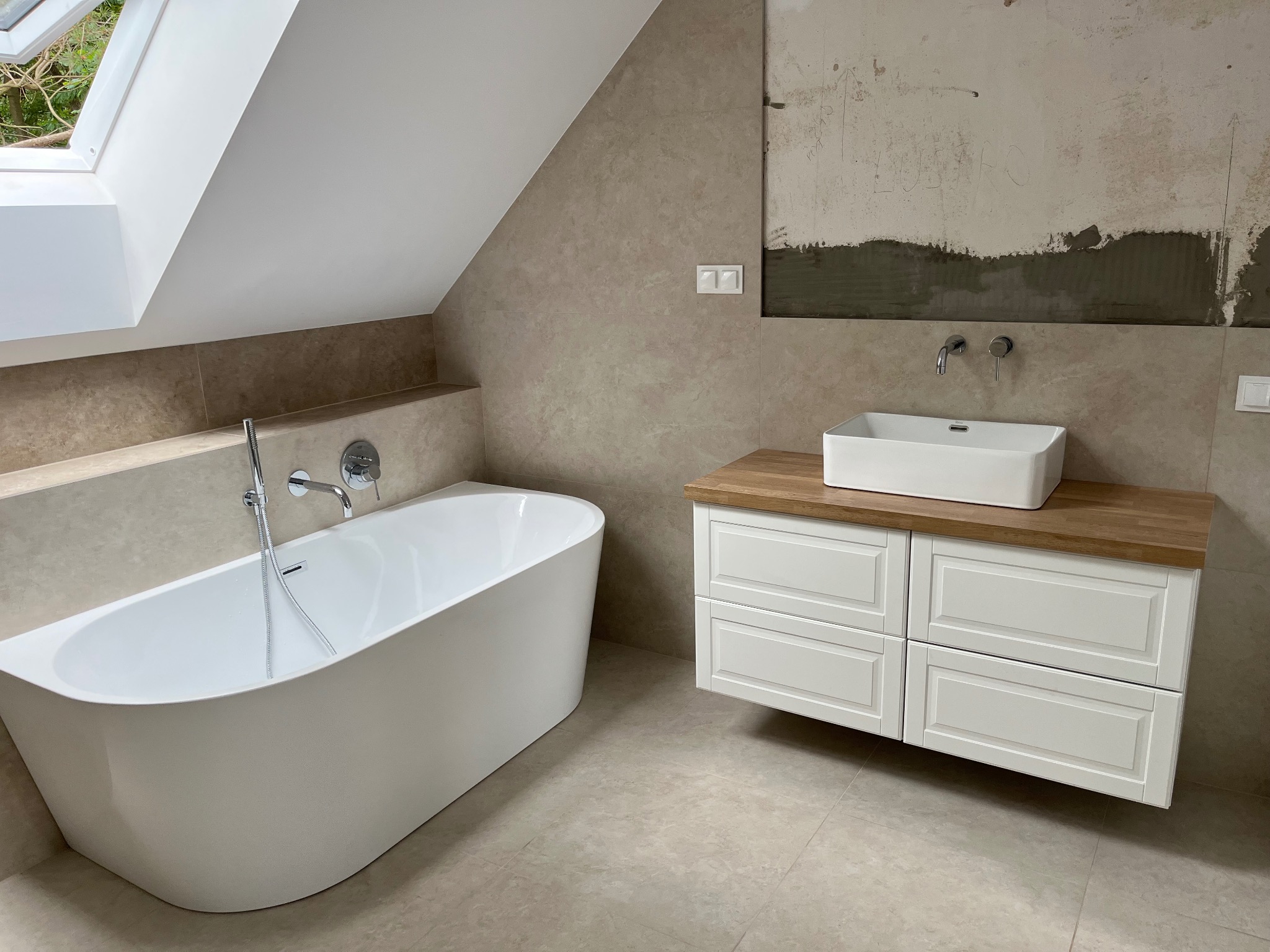 Nowoczesna łazienka na poddaszu z wolnostojącą wanną, umywalką nablatową na szafce z drewnianym blatem i beżowymi płytkami. Fragment ściany w trakcie wykończenia.