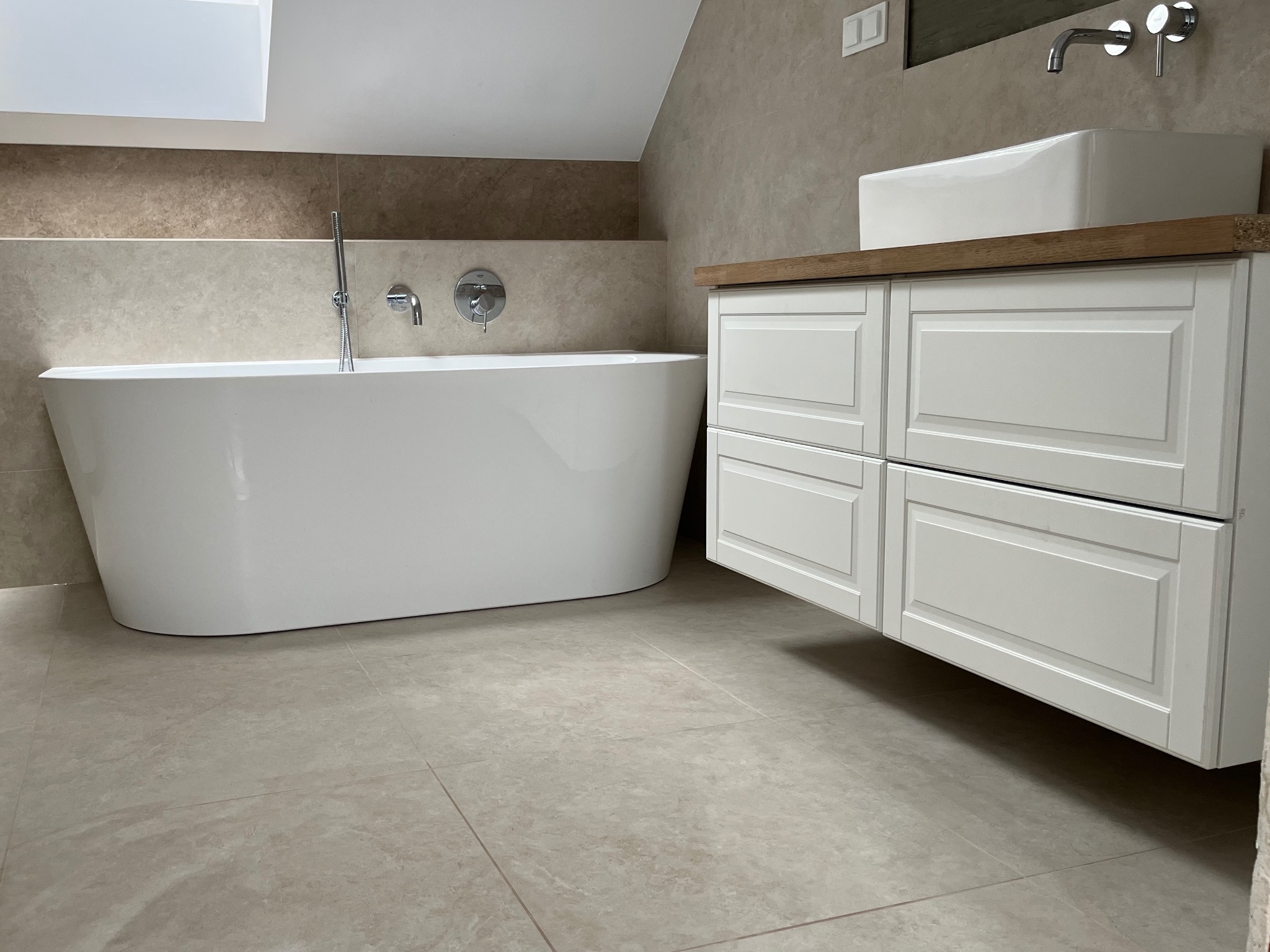 Nowoczesna łazienka w beżowej tonacji: wolnostojąca wanna, biała szafka z umywalką na drewnianym blacie, armatura w kolorze chromu i beżowe płytki podłogowe.