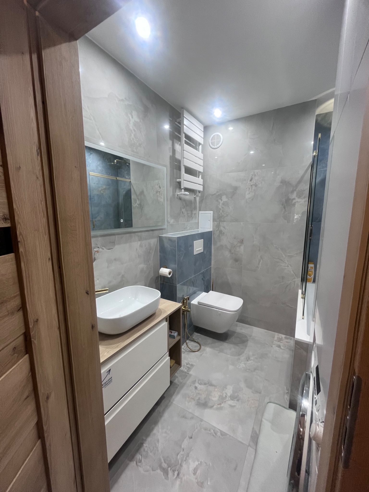 Stylowa łazienka w szarościach: marmurowe płytki, podwieszana toaleta, umywalka nablatowa z złotą armaturą i grzejnik drabinkowy. Nowoczesne oświetlenie.