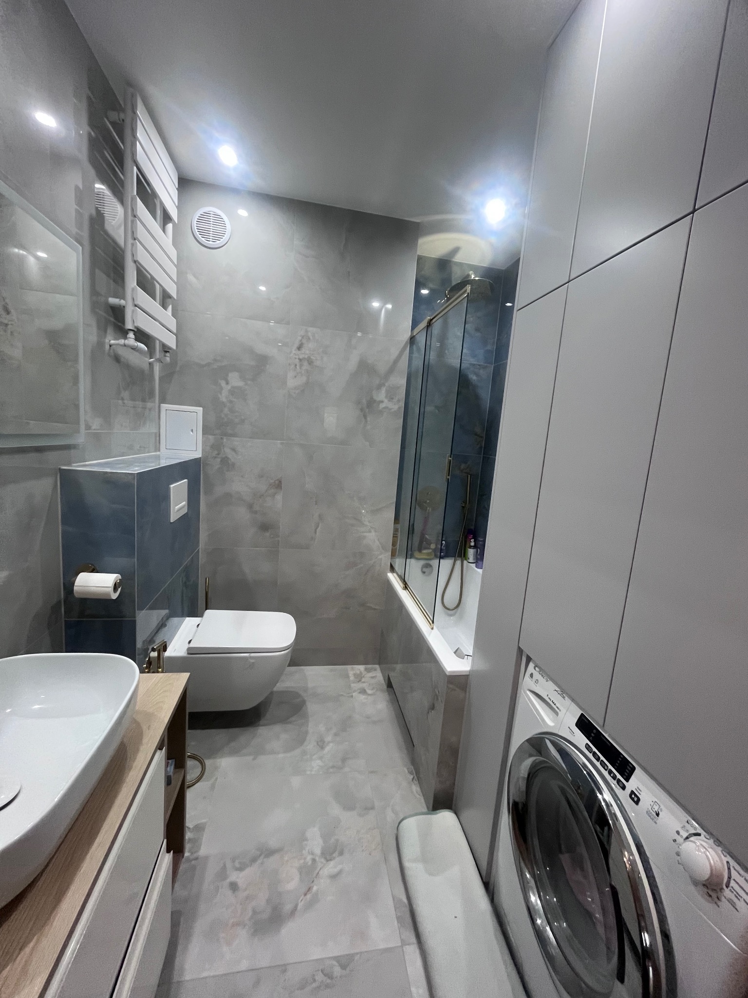 Nowoczesna łazienka w szarościach z marmurowymi płytkami, kabiną prysznicową nad wanną i pralką. Minimalistyczny design z podwieszaną toaletą i grzejnikiem drabinkowym.