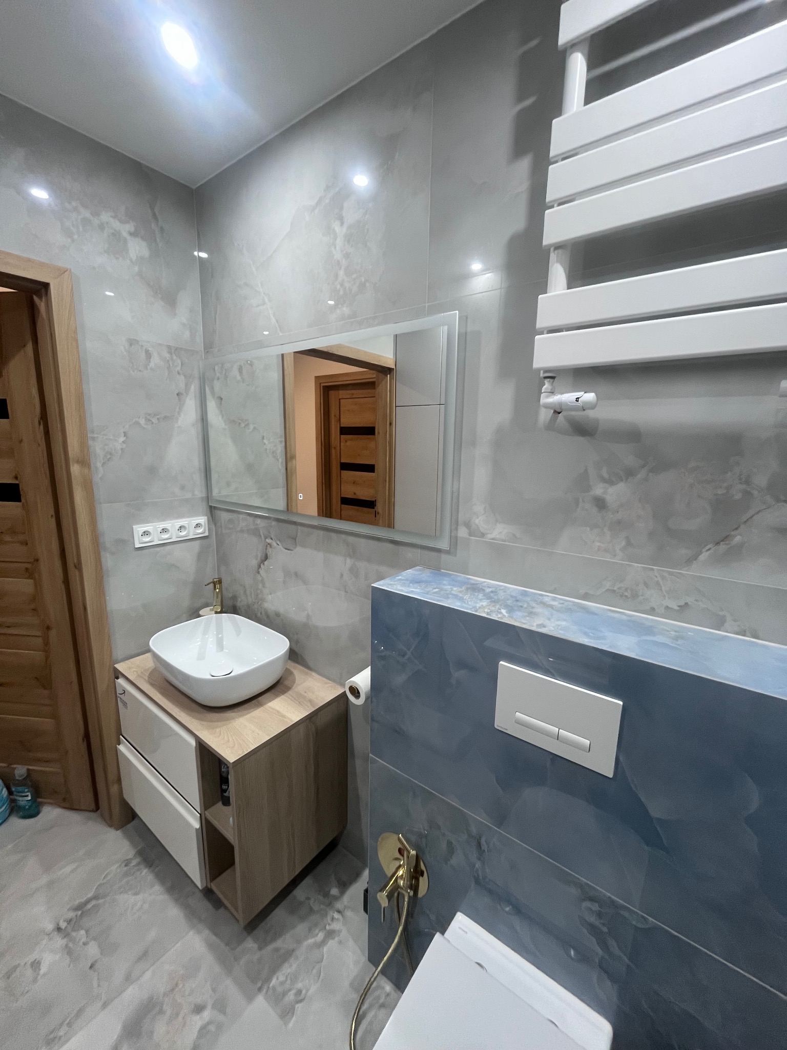 Nowoczesna łazienka z umywalką nablatową, lustrem z podświetleniem LED i grzejnikiem drabinkowym. Szare płytki imitujące marmur i akcenty w kolorze niebieskim.