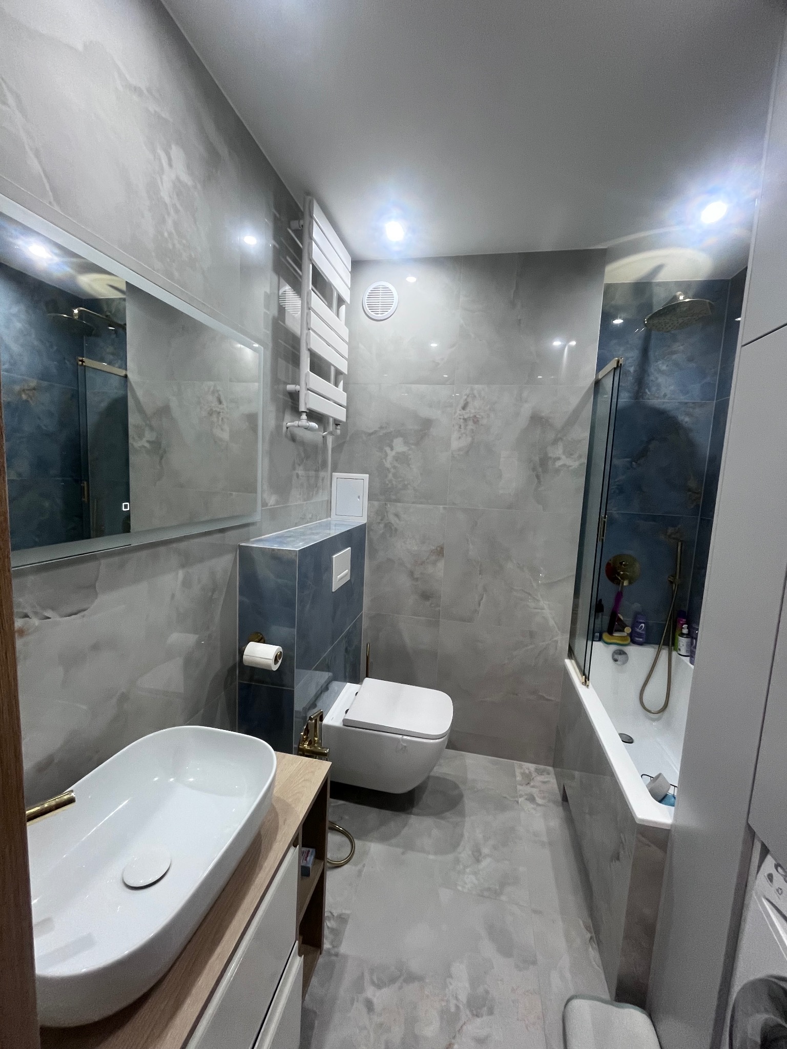 Nowoczesna łazienka z marmurowymi płytkami, owalną umywalką nablatową i kabiną prysznicową za szklaną ścianką. Grzejnik drabinkowy i oświetlenie punktowe.