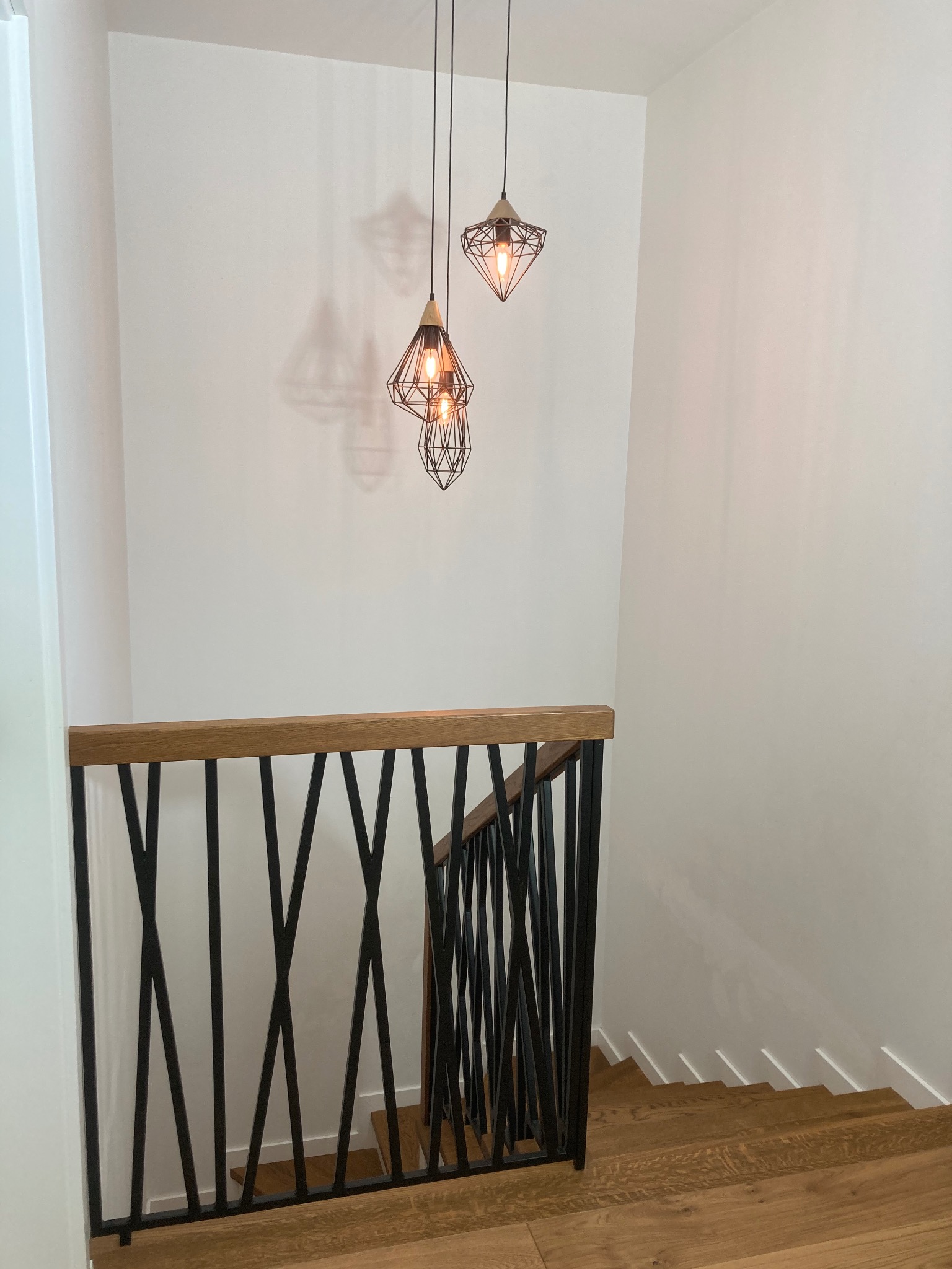 Nowoczesne schody z drewnianymi stopniami i czarną, metalową balustradą. Nad nimi wiszą trzy geometryczne lampy, rzucające cienie na białą ścianę.