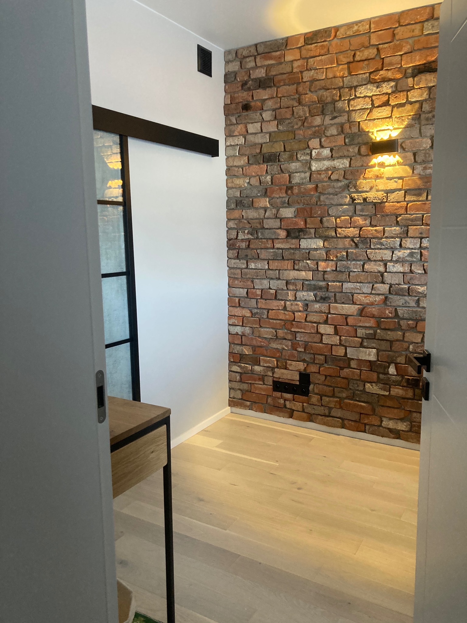 Wnętrze z ceglaną ścianą oświetloną kinkietami, jasną drewnianą podłogą i przesuwnymi drzwiami w czarnej ramie. Minimalistyczny design z akcentem industrialnym.