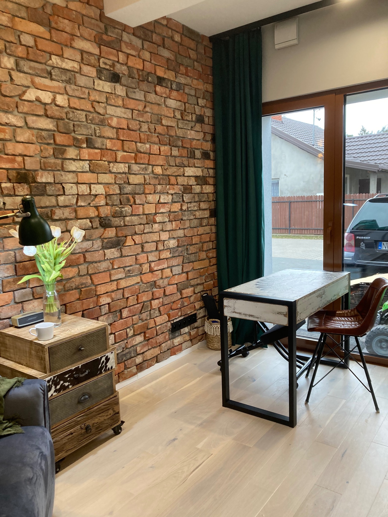 Loftowe wnętrze z ceglaną ścianą, drewnianą podłogą i designerskimi meblami. Widoczny stolik, krzesło i komoda w stylu vintage. Z okna widok na dom i samochód.
