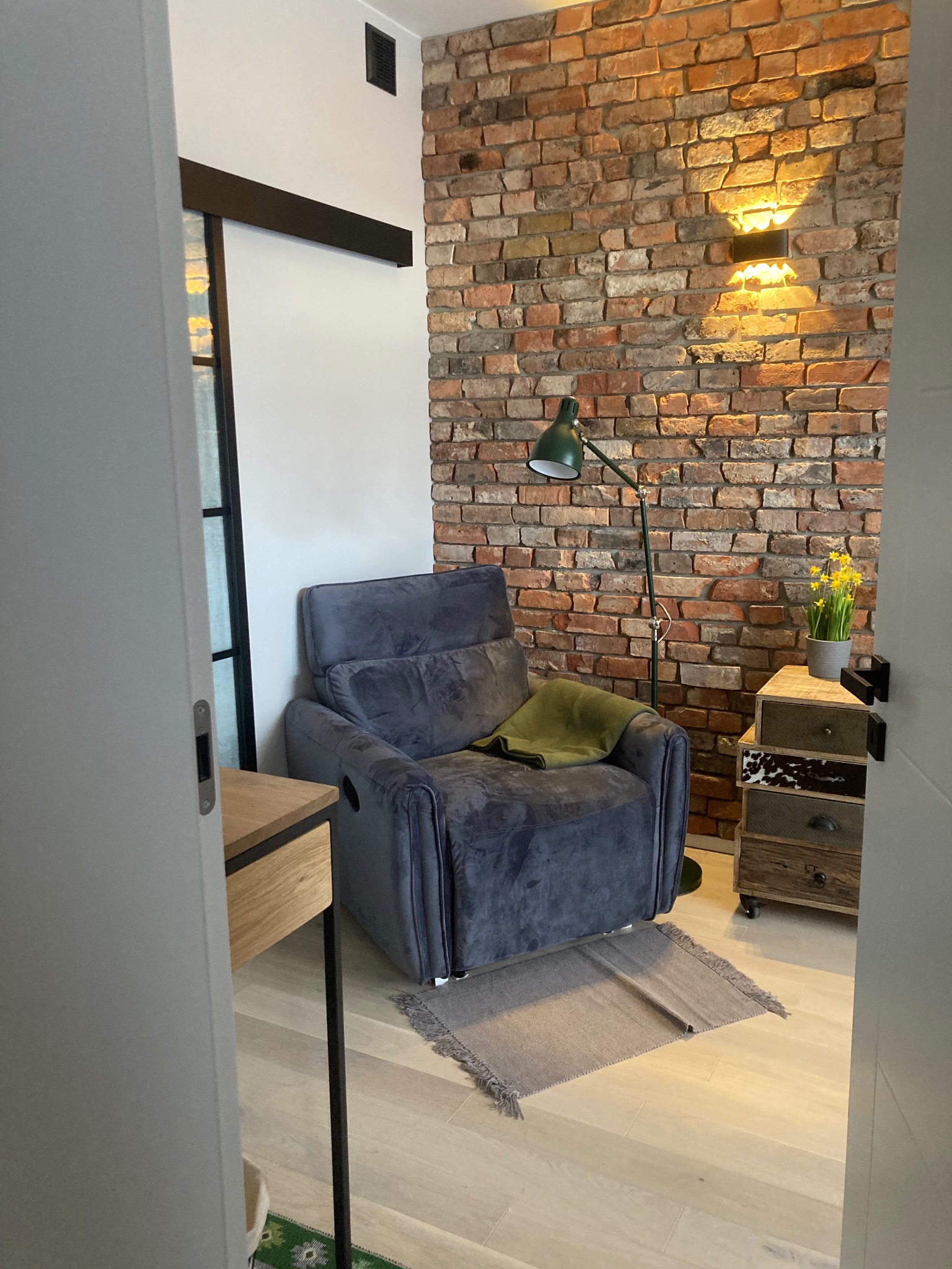 Stylowe wnętrze z ceglaną ścianą, szarym fotelem i lampą podłogową. Widoczna komoda z kwiatami i fragment drzwi przesuwnych. Przytulny kącik do relaksu.