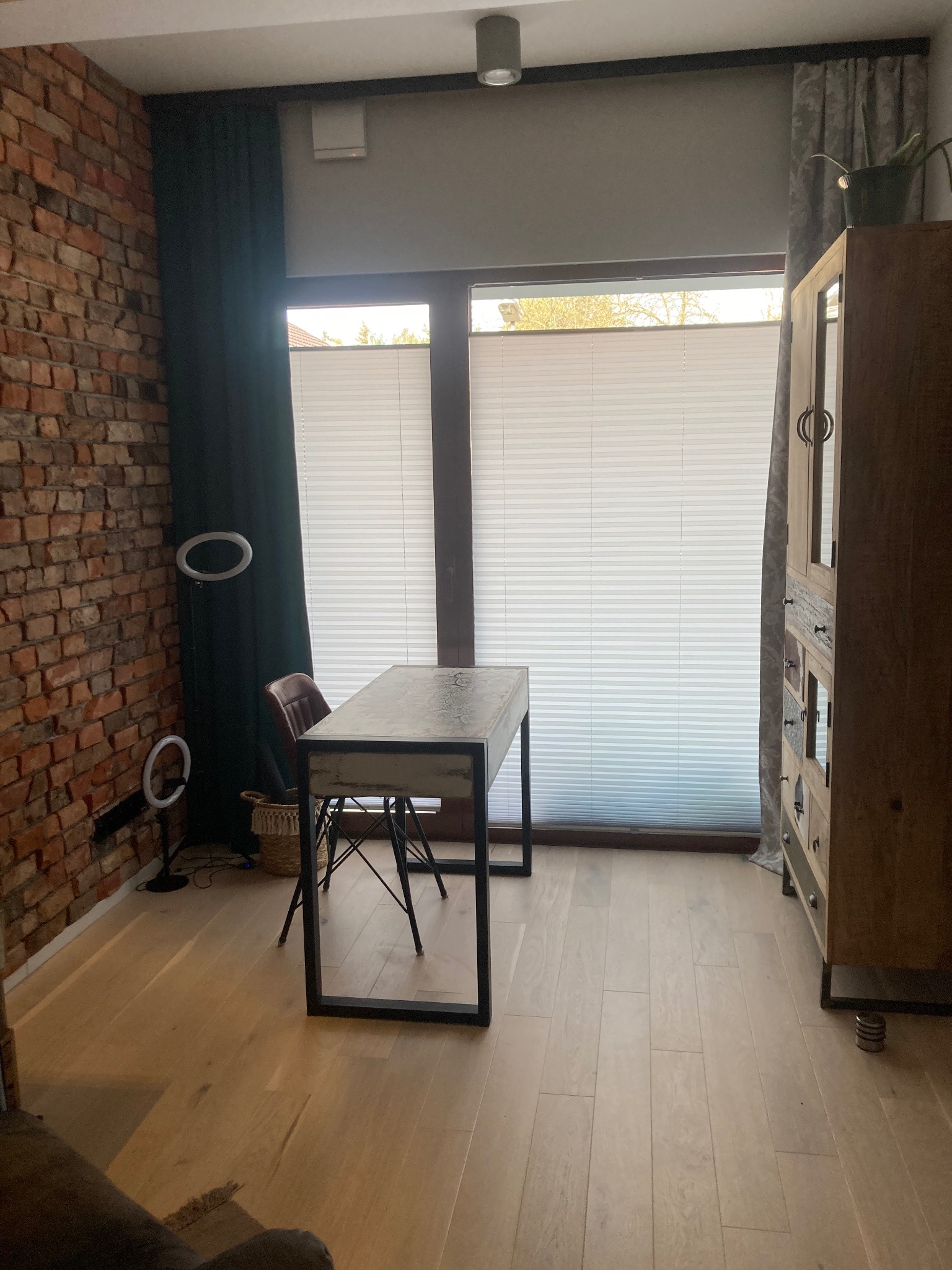 Loftowe wnętrze z ceglaną ścianą, drewnianą podłogą i biurkiem na tle okna z żaluzjami. Widoczne lampy pierścieniowe i drewniana komoda.