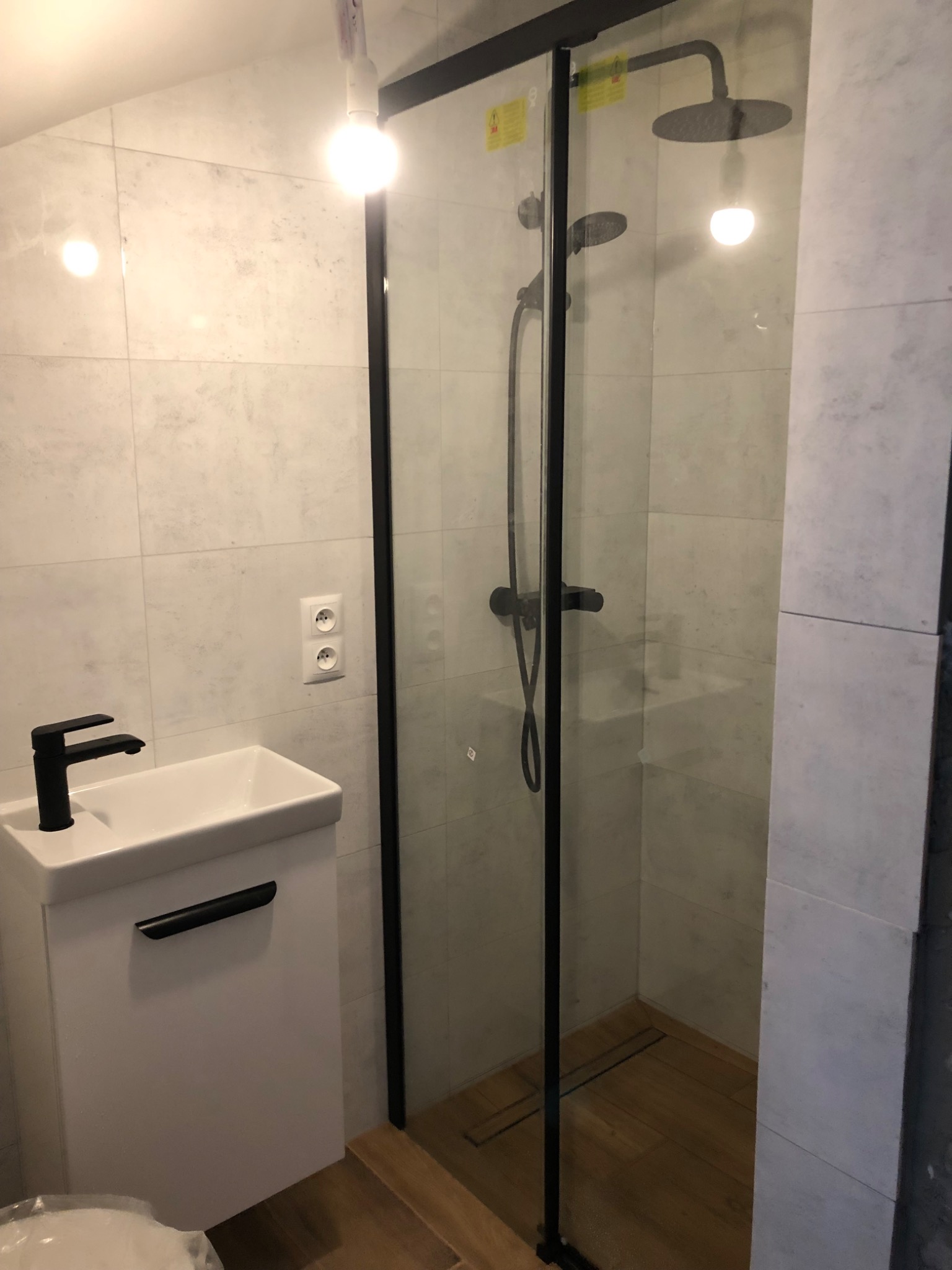 Nowoczesna łazienka z kabiną prysznicową typu walk-in z czarnymi ramami, białą umywalką z czarną armaturą i płytkami imitującymi kamień na ścianach.