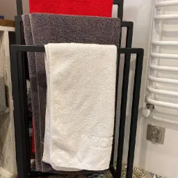 Czarny metalowy stojak na ręczniki z trzema ręcznikami w kolorach: czerwonym, szarym i białym. Ręczniki wiszą w łazience na tle płytek ceramicznych i grzejnika.