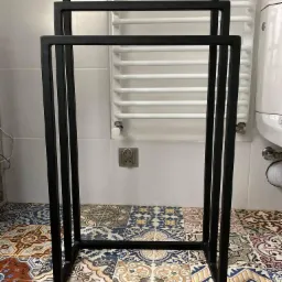 Czarny, metalowy stojak na ręczniki w łazience z kolorową, wzorzystą podłogą i białym grzejnikiem drabinkowym w tle.