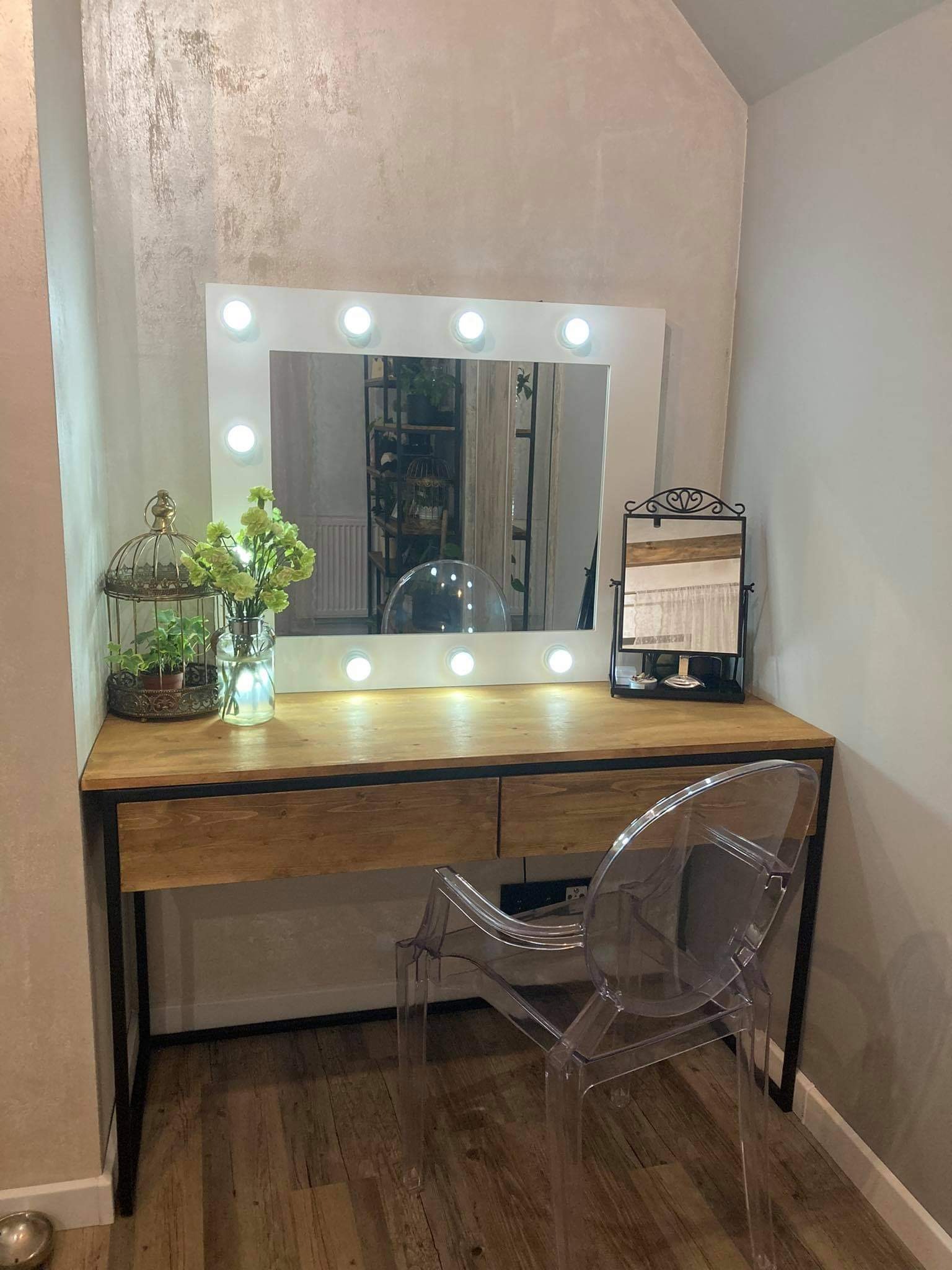 Drewniana toaletka z czarną metalową ramą, podświetlanym lustrem, przezroczystym krzesłem i dekoracjami w postaci klatki dla ptaków z rośliną i wazonu z kwiatami.