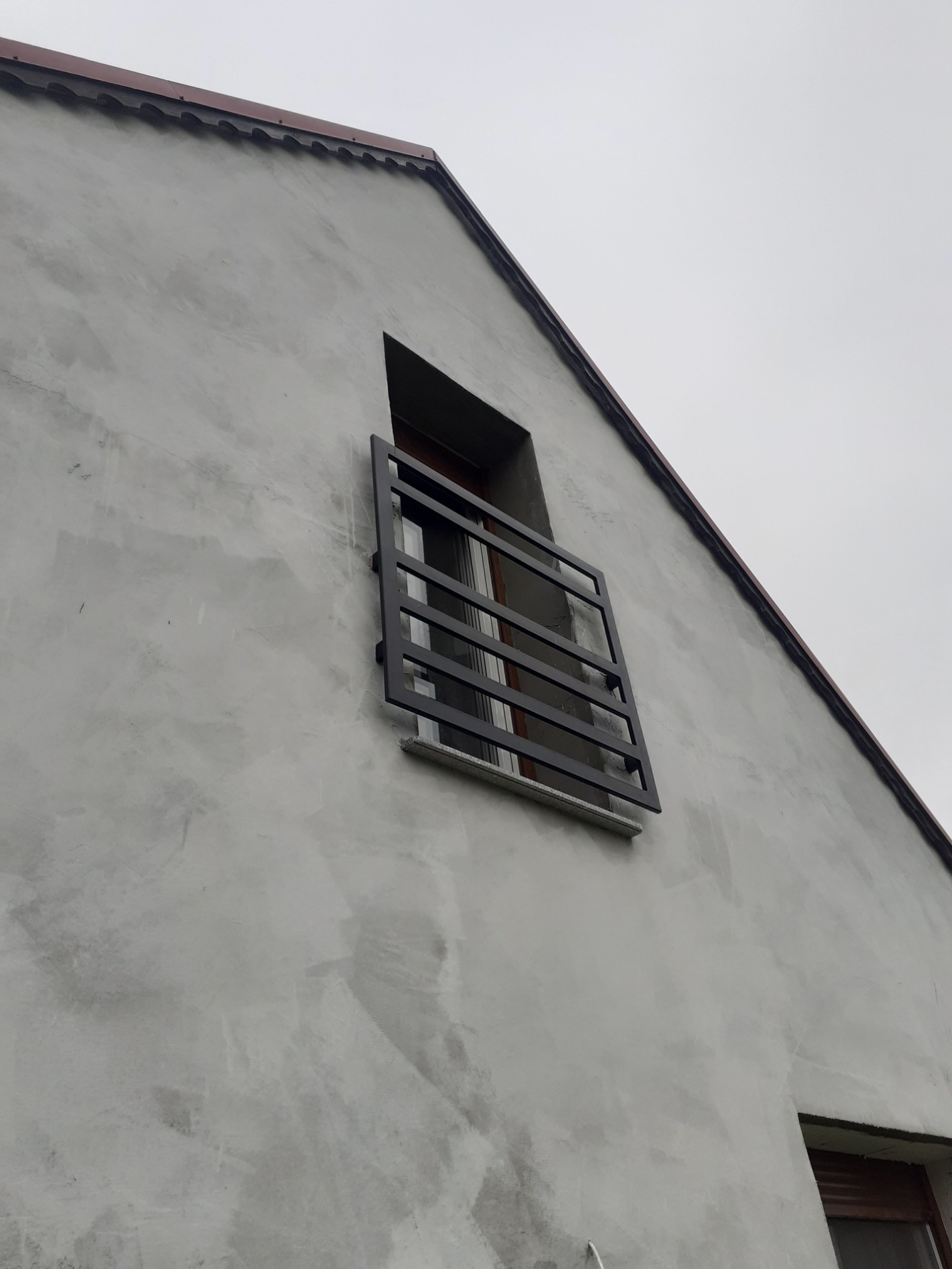 Zewnętrzna krata okienna z ciemnego metalu, zamontowana na surowej, szarej ścianie budynku z widocznym oknem w tle.