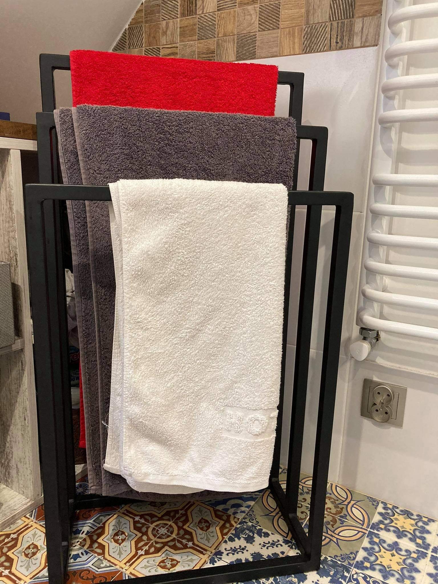 Czarny metalowy stojak na ręczniki z trzema ręcznikami w kolorach: czerwonym, szarym i białym. Ręczniki wiszą w łazience na tle płytek ceramicznych i grzejnika.