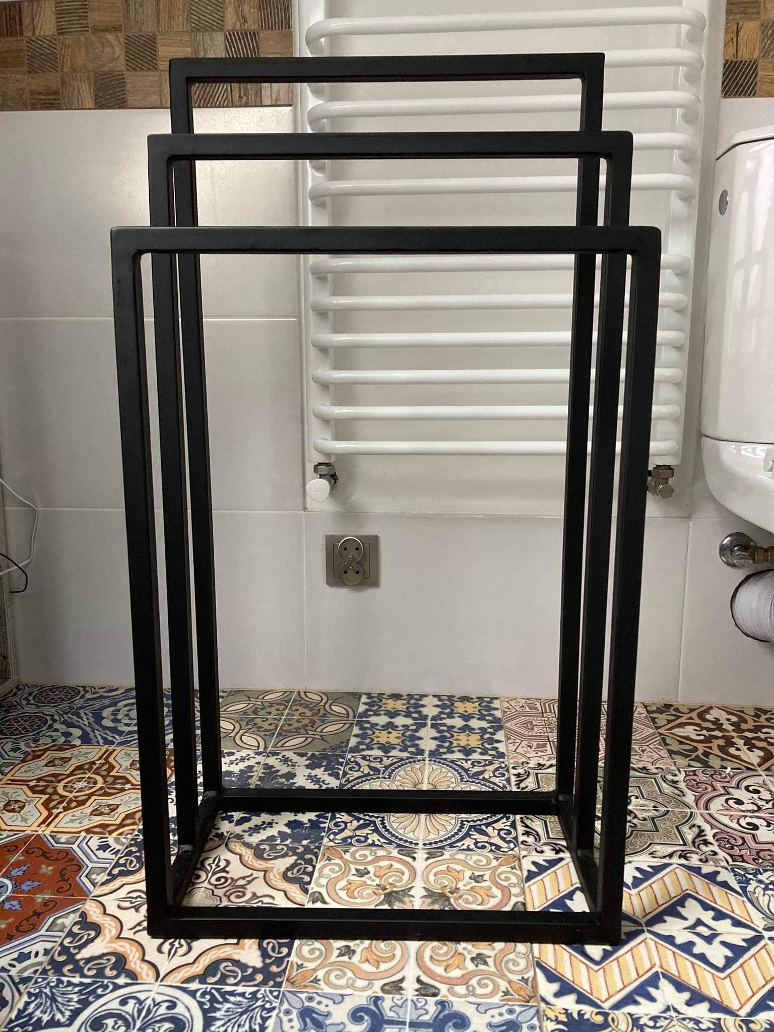 Czarny, metalowy stojak na ręczniki w łazience z kolorową, wzorzystą podłogą i białym grzejnikiem drabinkowym w tle.