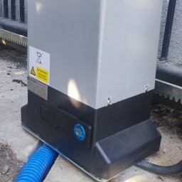 P.H.U TECH-MIKI - Szary automat bramy przesuwnej marki ElektroBM z niebieską rurą kablową, zamontowany na betonowej podstawie przy metalowym ogrodzeniu.