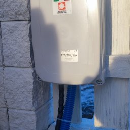 P.H.U TECH-MIKI - Szara skrzynka sterownicza Roger Technology B70/2ML/BOX z niebieskim i czarnym przewodem zamontowana na białym ogrodzeniu i kamiennym słupku.