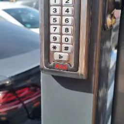 Szary panel domofonu ATLO z klawiaturą numeryczną na słupku bramy wjazdowej. W tle fragment samochodu i budynku.