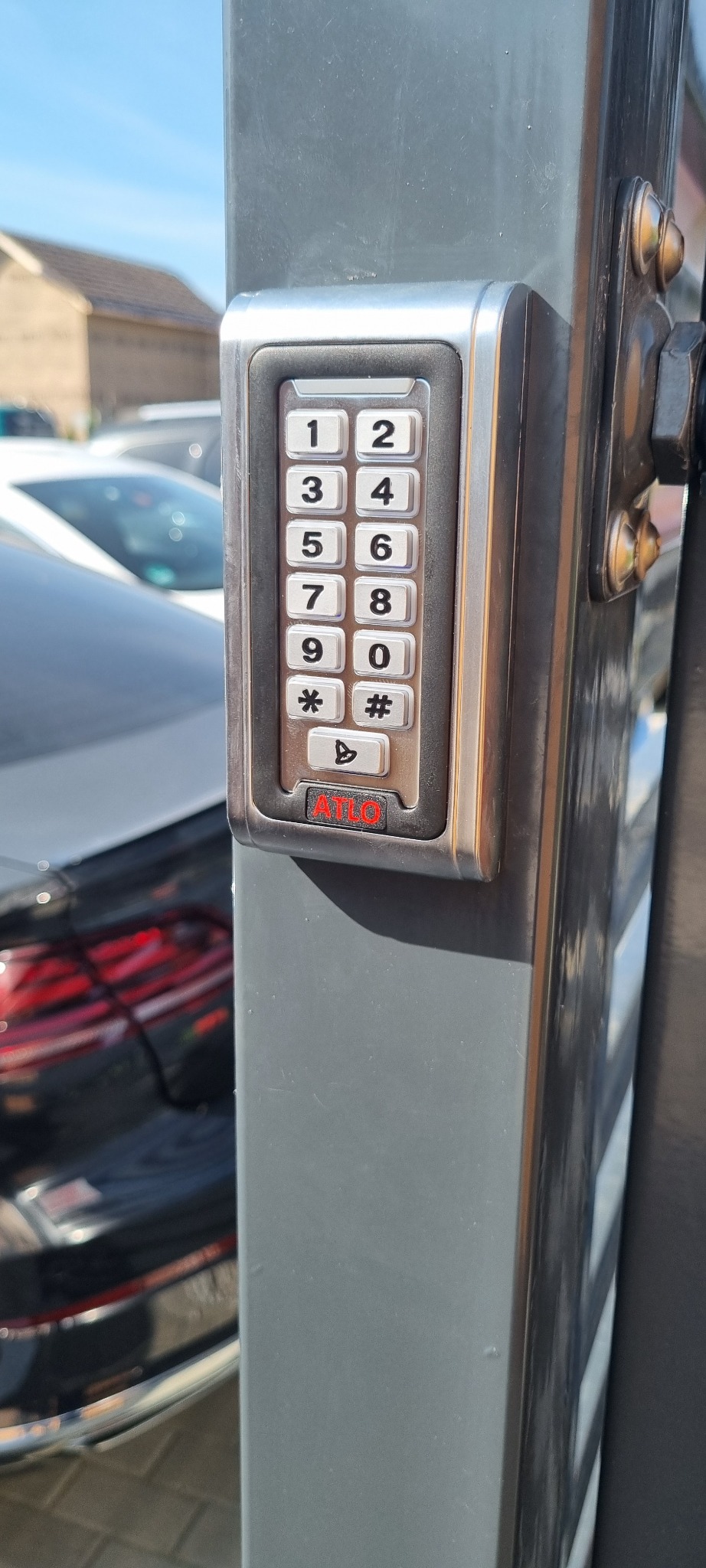Szary panel domofonu ATLO z klawiaturą numeryczną na słupku bramy wjazdowej. W tle fragment samochodu i budynku.