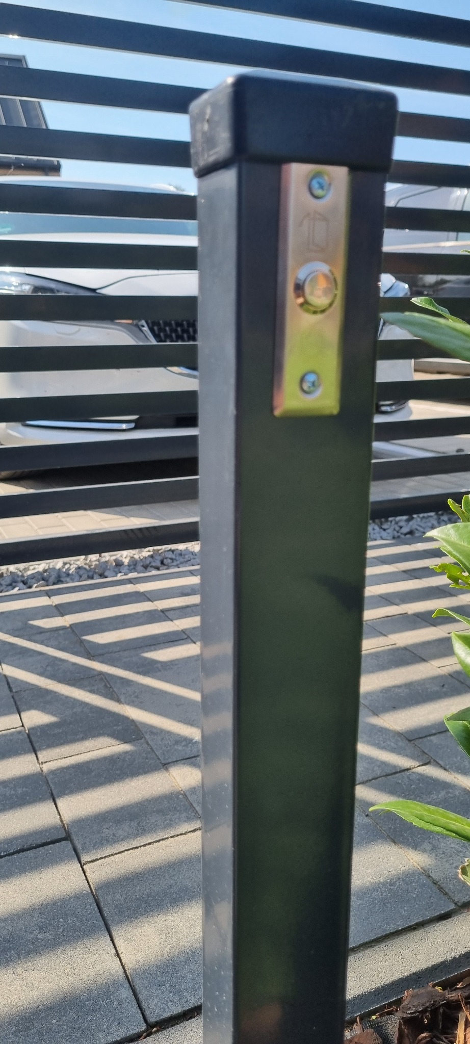 Metalowy słupek z przyciskiem domofonu na tle nowoczesnej, poziomej bramy i kostki brukowej. Widoczne elementy roślinności i fragment samochodu.